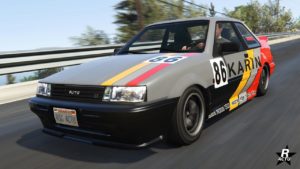 Image présentant le véhicule Karin Futo de l'avant. La voiture de GTA Online est peinte en grise, sa carrosserie est recouverte de décalcomanie de course automobile où des bandes rouges et jaunes sont présentes, ainsi que le numéro de course "86" sur les portes latérales.