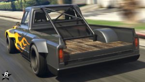 Photo d’un pickup vue de derrière avec une carrosserie grise et un design de flamme distinctif sur le côté. Le lit du camion est bordé de planches de bois, et il y a une structure de barre anti-roulis derrière la cabine. Le véhicule est représenté en mouvement sur ce qui semble être une route avec des environs flous, suggérant la vitesse.