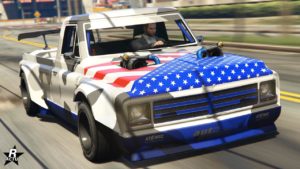 Image d’un véhicule du jeu vidéo GTA Online, spécifiquement le Declasse Yosemite Drift. Il est représenté avec une livrée blanche, bleue et rouge comportant des étoiles et des rayures, ressemblant au drapeau américain. Le véhicule a une carrosserie modifiée avec des éléments aérodynamiques ajoutés comme un splitter avant et un aileron arrière, indiquant qu’il est conçu pour le drift. Le capot est relevé à l’arrière, suggérant une modification du moteur ou une amélioration du système de refroidissement. Il y a deux grands tuyaux d’échappement qui sortent des ailes avant. Le conducteur est visible à l’intérieur de la voiture avec les deux mains sur le volant, portant une tenue décontractée.