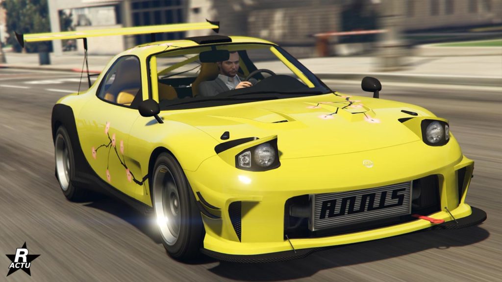 Cette image représente une voiture de sport, l'Annis ZR350 dans le jeu vidéo GTA Online. La voiture est jaune vif avec des décalcomanies de fleurs roses et blanches sur le côté. Elle a un design agressif avec un large pare-chocs avant, des phares proéminents et un capot plat. Un aileron arrière noir est visible à l’arrière du véhicule. L’environnement autour de la voiture semble est une rue en ville pendant la journée avec des bâtiments en arrière-plan et un ciel clair.