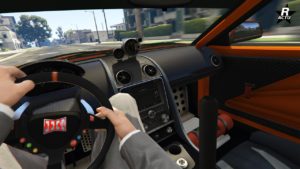 Cette image est une capture d’écran d’un jeu vidéo montrant l’intérieur d’une voiture, la Dinka Jester RR dans GTA Online. La vue est à la première personne depuis le siège du conducteur. Le conducteur tient le volant. L’intérieur de la voiture est détaillé, avec des couleurs dominantes noir et orange. Divers contrôles et affichages sont visibles, y compris des bouches d’aération, des boutons et une interface tactile centrale qui semble être éteinte ou ne pas afficher d’informations. Une partie de l’environnement extérieur virtuel est visible à travers le pare-brise et la fenêtre latérale qui donne sur une zone urbaine avec des bâtiments et des arbres visibles à l’extérieur.