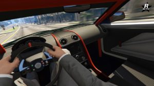 Cette image montre l’intérieur d’une voiture, il s'agit de la sportive japonaise Jester RR du point de vue du conducteur dans le jeu vidéo GTA Online. Le conducteur tient un volant noir avec les deux mains ; il porte un costume gris et une chemise blanche. Le tableau de bord est bien en évidence, avec trois bouches d’aération circulaires et une garniture orange qui le souligne. On aperçoit la route et les environs à travers le pare-brise, indiquant que la voiture est en mouvement dans une rue de la ville en plein jour.