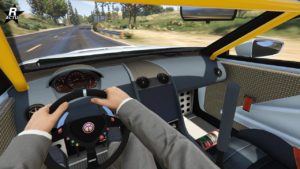 Cette image représente une vue à la première personne de l’intérieur d’une voiture du jeu vidéo Grand Theft Auto Online. Le véhicule est l'Emperor Vectre. Les mains du conducteur sont visibles sur le volant, il porte un costume gris. Le volant a plusieurs boutons de différentes couleurs et un logo au centre. Il y a une vue claire du compteur de vitesse de la voiture et d’autres jauges derrière le volant. L’intérieur comprend des détails comme des bouches d’aération, des matériaux texturés sur les portes et le tableau de bord, et des ceintures de sécurité. Une cage de sécurité jaune est visible au-dessus et sur le côté, indiquant qu’il s'agit d’un modèle de voiture de sport. À l’extérieur du pare-brise, il y a une scène floue d’une route entourée de verdure sous une lumière du jour vive.