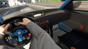 Vue intérieure d’une voiture de course Dinka Kanjo SJ dans le jeu vidéo GTA Online, du point de vue du conducteur. Les mains du conducteur sont sur un volant bleu et noir avec divers boutons et commandes. Le tableau de bord comprend un ensemble de commutateurs et de boutons de différentes couleurs et symboles, indiquant des fonctionnalités complexes. Un affichage numérique est intégré dans le tableau de bord, bien que ses lectures ne soient pas claires. Le panneau de porte intérieur est visible avec un arceau de sécurité et une poignée de tirage orange pour ouvrir la porte. Un rétroviseur latéral donne un aperçu de la route et des environs à l’extérieur de la voiture. Le conducteur porte un costume gris.