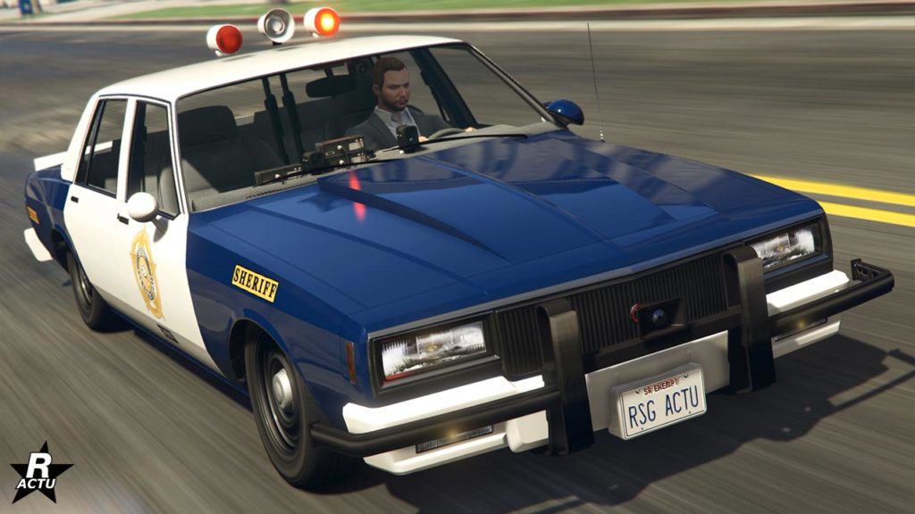 Vue avant de la voiture de police Declasse Impaler LX Cruiser dans le jeu vidéo GTA Online. La voiture est de couleur bleu foncé et blanc, typique des véhicules de police. L'insigne de shérif en jaune se trouve sur les portes latérales. Le véhicule roule à vive allure sur une route de Los Santos, avec les gyrophares plutôt anciens allumés.