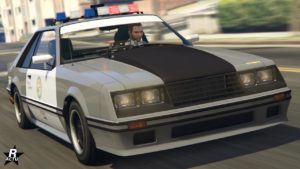 La voiture de flic Vapid Dominator FX Interceptor vue de devant. Elle a un duo de couleurs gris clair et blanc, ainsi que le logo du LSPD sur les portes. Le centre du capot ressort de quelques centimètres vers le haut tout en étant en carbone. Sur le toit, des gyrophares typiques des années 1980 sont allumés.