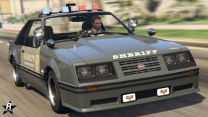 Vue avant du véhicule Vapid Dominator FX Interceptor dans GTA Online. Il s'agit d'une voiture de police des années 1980 qui s'inspire d'une Ford Mustang de troisième génération. La carrosserie est de couleur gris et beige, le logo du shérif se trouve un peu partout sur le véhicule.