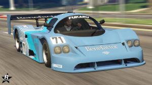 Vue avant d’une voiture de course bleu clair, la ‘Benefactor LM87’, dans le jeu vidéo GTA Online. La voiture porte le numéro 71 et présente des marques proéminentes comme ‘Fukaru’, ‘Xero’ et ‘Terroil’ sur le devant et les côtés. Elle est équipée d’un grand aileron arrière, et d’une paire de phares circulaires. Le véhicule est représenté en mouvement sur une route de Los Santos, comme l’indique le flou de mouvement autour du véhicule.