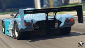 Vue arrière d’une voiture de course bleu clair, la ‘Benefactor LM87’, dans le jeu vidéo GTA Online. Le véhicule présente un aileron arrière proéminent, des doubles tuyaux d’échappement et des feux arrière circulaires distinctifs. La plaque d’immatriculation indique ‘ RSG ACTU’. Le cadre de l'image est une journée ensoleillée dans une rue de la ville avec des palmiers en arrière-plan.