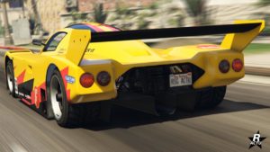 Vue arrière d’une voiture de course jaune, la ‘Benefactor LM87’, dans le jeu vidéo GTA Online. La voiture présente un grand aileron arrière noir. Le véhicule est représenté en mouvement sur ce qui est une rue de Los Santos avec un arrière-plan flou suggérant la vitesse. La voiture présente des décalcomanies de course, incluant le numéro ‘87’, et a des feux arrière rouges circulaires, des tuyaux d’échappement doubles, et une plaque d’immatriculation qui indique ‘RSG ACTU’.