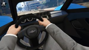 Vue intérieure de la voiture Dewbauchee Vagner dans le jeu vidéo GTA Online. L'image est du point de vue du conducteur. Les mains du conducteur sont visibles sur un volant noir avec l'emblème du constructeur Dewbauchee au centre. Le tableau de bord derrière le volant contient diverses jauges et cadrans. Le design intérieur présente des textures en fibre de carbone et des accents métalliques élégants sur le tableau de bord. À travers le pare-brise, une scène de rue urbaine avec des bâtiments, des feux de circulation et des arbres verdoyants est visible.