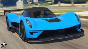 Vue de face d’une voiture de sport Dewbauchee Vagner bleu vif du jeu vidéo GTA Online. La voiture présente un design élégant avec des angles vifs et des caractéristiques aérodynamiques. Elle est équipée d’un aileron arrière noir proéminent, de phares LED distinctifs et d’une grande grille noire sur le pare-chocs avant. L’image est prise sur une route dans le jeu, avec un flou de mouvement en arrière-plan indiquant la vitesse de la voiture.