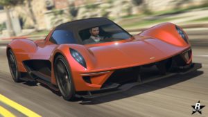 Vue avant d’une Dewbauchee Vagner orange du jeu vidéo Grand Theft Auto Online. La voiture est en mouvement sur une route du jeu. Le design du véhicule présente des lignes aérodynamiques élégantes, avec des ailes avant proéminentes et une position basse qui suggère une grande vitesse et une ingénierie avancée typique des supercars.