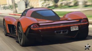 Vue arrière d’une voiture de sport moderne Dewbauchee Vagner orange avec des détails noirs distinctifs dans le jeu vidéo GTA Online. La voiture est en mouvement dans une rue de la ville, comme l’indique l’arrière-plan flou suggérant la vitesse, et elle dispose d'un petit aileron plat. La plaque d’immatriculation indique ‘RSG ACTU’.