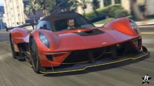 Vue avant d’une voiture de sport Dewbauchee Vagner rouge avec des accents noirs du jeu vidéo GTA Online. La voiture est en mouvement sur une rue de la ville, avec des environs flous suggérant une grande vitesse. Le design de la voiture présente un profil bas, une carrosserie aérodynamique, de grandes prises d’air à l’avant et un aileron arrière distinctif pour la portance.