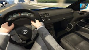 L’image montre l’intérieur d’un véhicule du point de vue du conducteur. Les éléments visibles comprennent un volant avec un logo au centre, qui est celui d’Annis, une marque de voiture fictive du jeu vidéo Grand Theft Auto. Le tableau de bord contient diverses jauges et indicateurs, dont un compteur de vitesse et un compte-tours. La console centrale a plusieurs boutons et une interface de système audio. Le pare-brise offre une vue sur une route floue devant, suggérant un mouvement.