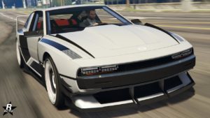 L’image montre une vue avant d’une voiture Bollokan Envisage grise du jeu vidéo GTA Online. Le véhicule présente un design de garniture noire distinctif avec des angles vifs et un capot plat, ce qui lui donne un aspect sportif. La voiture a des phares rectangulaires en LED, une grille noire avec le logo Bollokan au centre, et un pare-chocs avant agressif avec de grandes prises d’air. On peut également voir un personnage à l’intérieur de la voiture à travers le pare-brise.