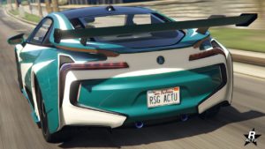 L’image montre la vue arrière d’une voiture de sport vert pétrole avec un grand aileron arrière noir. La voiture est l'Ubermacht Niobe et présente des lignes blanches distinctives accentuant son design aérodynamique, et elle arbore un emblème du fabricant au centre, au-dessus de la plaque d’immatriculation, qui porte l’inscription “RSG ACTU”. Le véhicule se trouve sur ce qui semble être une route, avec une végétation floue en arrière-plan, suggérant le mouvement.