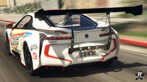 L’image montre une voiture Ubermacht Niobe blanche provenant du jeu vidéo GTA Online, vue de l’arrière. La voiture est ornée de divers autocollants de course et de logos de sponsors, avec un aileron arrière imposant pour l’aérodynamisme. La plaque d’immatriculation porte l’inscription “RSG ACTU”.