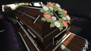 Image de l'arrière du corbillard, un cercueil en bois se trouve à l'arrière de la voiture, une couronne de fleurs se trouve posée dessus.
