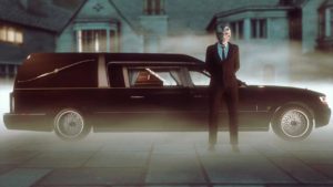 Image présentant le Chariot Corbillard Romero d'une vue latérale. Le véhicule est de couleur noire, se trouve entouré d'un brouillard et un personnage de GTA Online avec un masque d'halloween se trouve devant la voiture.