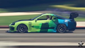 Vue latérale de la voiture japonaise Dinka Jester Classique dans le jeu vidéo GTA Online. Le véhicule roule rapidement comme en témoigne le flou de mouvement autour de la sportive. Sa carrosserie est peinte en vert, un grand aileron de course en carbone se trouve à l'arrière, et une décalcomanie géométrique de plusieurs nuances de bleues est présente sur la voiture.