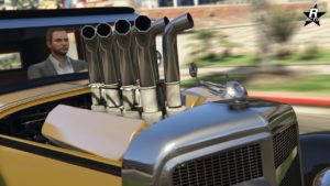 Vue proche du capot de la voiture Albany Roosevelt Valor dans GTA Online, des tuyaux de refroidissements du moteur dépassent du capot. L'emblème ailé du constructeur britannique Enus se trouve au-dessus de la calandre.