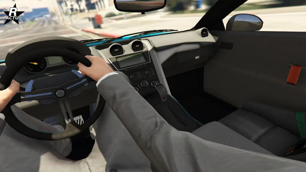 L'intérieur du véhicule 10F Large / Widebody du point du vue du conducteur lors de sa conduite dans le jeu vidéo GTA Online. Les portières sont recouvertes de fibres avec une languette rouge en guise de poignée. Le tableau de bord est gris, la console centrale à des coutures bleues pour finitions et derrière le volant se trouve deux compteurs analogiques et un petit écran entre les deux.