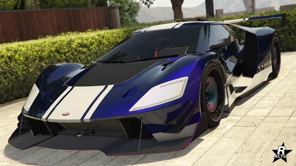 Vue avant de la supercar Vapid FMJ MK V, photographiée dans l’allée d’un manoir de GTA Online. Le véhicule arbore une teinte bleu foncé, rehaussée de reflets bleus plus clairs. Une large bande blanche, en forme de décalcomanie, traverse verticalement la carrosserie, du capot jusqu’au toit. Le haut du capot, quant à lui, est en carbone. Un splitter aérodynamique en carbone est fixé sous le pare-chocs avant qui est flanqué de deux moustaches en carbone de parte d'autres. Sur le côté droit du véhicule, une seconde bande blanche se trouve entre les deux roues, au niveau du bas de caisse.