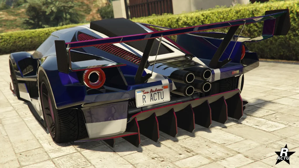 Image présentant l’arrière de la supercar Vapid FMJ MK V, issue du jeu vidéo Grand Theft Auto Online. Le véhicule arbore une teinte bleu foncé, rehaussée de reflets bleus clairs. Des finitions aux nuances de rouge et de rose soulignent les contours du diffuseur arrière en forme de râteau, situé sous le pare-chocs. Ces mêmes accents colorés ornent également le grand aileron de course et les lamelles horizontales entre le capot moteur en carbone, qui relie le toit aux quatre pots d’échappement centraux.