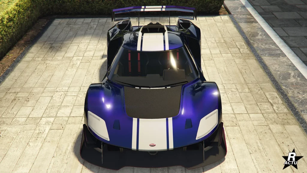 Vue de face de la supercar américaine Vapid FMJ MK V, dans la cour d’un manoir de GTA Online. Le véhicule arbore une peinture bleue foncée avec des reflets de bleus plus clairs. Une décalcomanie, sous la forme d’une bande blanche assez large, traverse le centre de la carrosserie de la voiture de manière verticale. Sur le toit du bolide, se trouve une entrée d’air en carbone à trois ouvertures.