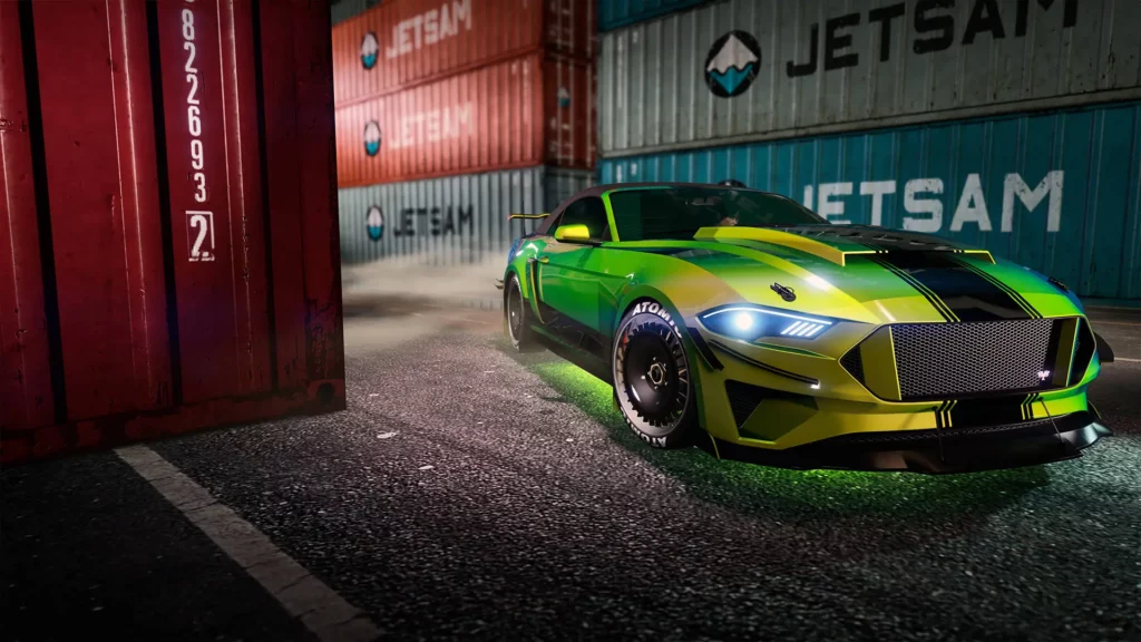 Image présentant la voiture Vapid Dominator GT avec une vue avant de celle-ci, dans le quartier portuaire de GTA Online, avec des conteneurs autour du véhicule, peint avec une couleur caméléon vert et disposant d'un motif d'une bande large noire verticale sur sa carrosserie.