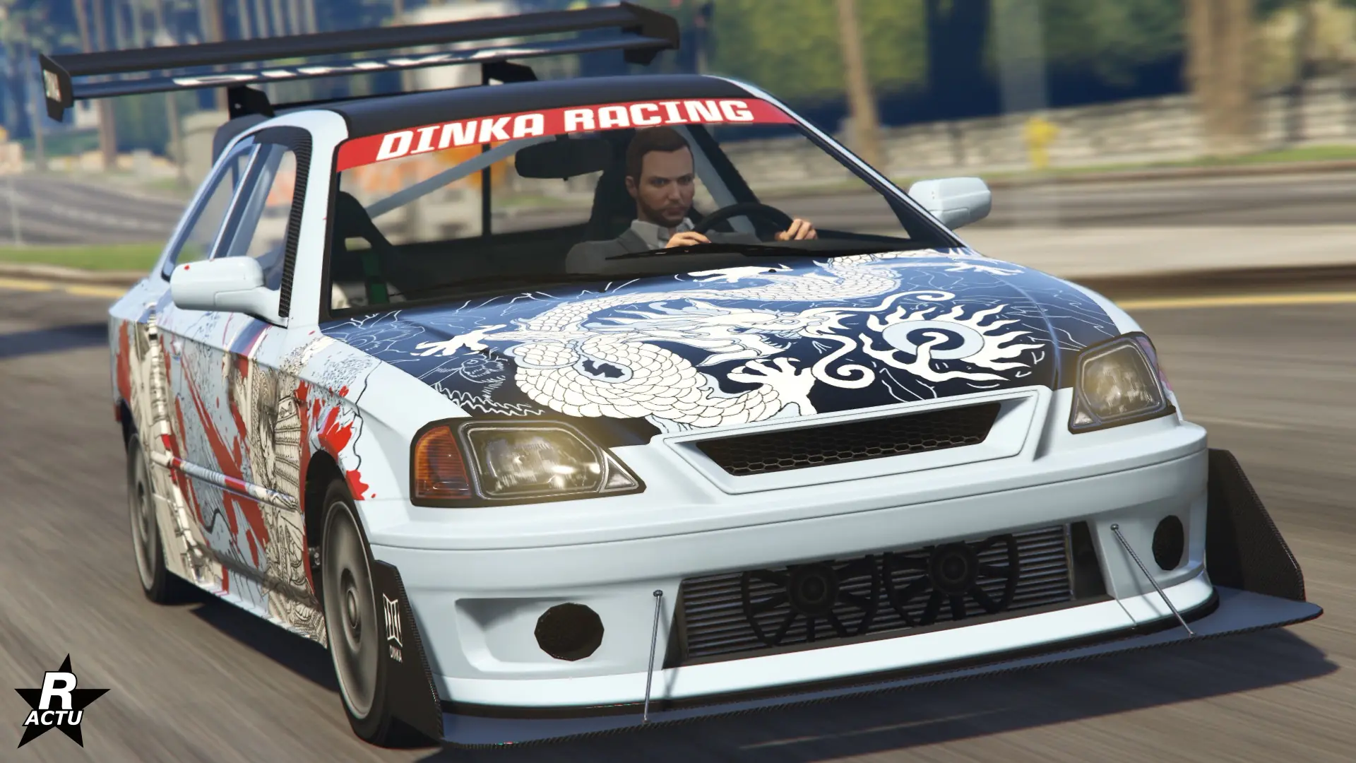 Vue avant de la voiture japonaise Dinka Blista Kanjo circulant sur une route ensoleillée dans le jeu vidéo GTA Online de Rockstar Games. Le véhicule à sa carrosserie peinte en blanc et dispose du motif rare « Samurai de rue ». Ce motif ajoute une décalcomanie d'un samouraï sur les côtés latéraux du bolide et la représentation d'un dragon blanc sur le capot de couleur noir. Le pare-chocs avant dispose de l'option ajoutant un grand séparateur aérodynamique noir et du radiateur d'air apparent. au niveau du pare-brise avant, se trouve un pare-soleil rouge avec l'inscription « Dinka Racing ».